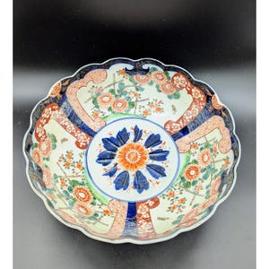 May include: A decorative ceramic bowl with a scalloped edge. The bowl features a central floral design in orange and blue, surrounded by a cream-colored background with orange and green floral patterns. The rim is detailed with a dark blue and red design.