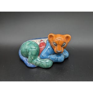 May include: A ceramic figurine of a reclining bear. The bear has an orange head and is resting on a blue base. The base has green and white accents. The figurine is a decorative item.