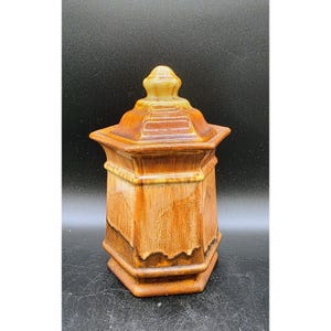 May include: A decorative, hexagonal ceramic jar with a lid. The jar is primarily brown with lighter yellow and tan accents. The lid has a small, rounded finial. The jar has a glossy finish and is likely vintage.