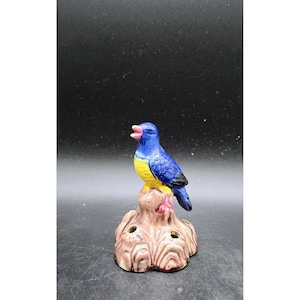 May include: A ceramic bird figurine with blue, yellow, and pink coloring. The bird is perched on a brown tree stump base with two holes. The bird has its beak open.