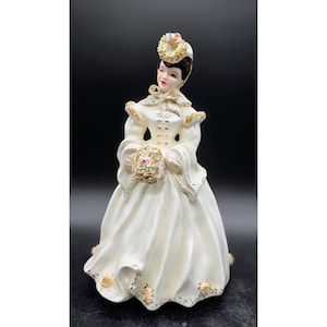 May include: A vintage porcelain figurine of a woman in a long, cream-colored dress with gold accents. The figurine holds a bouquet and wears a matching hat. The dress has a full skirt and long sleeves, with gold trim on the edges.