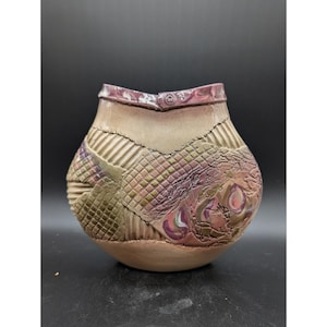 May include: A handcrafted ceramic vase with a unique patchwork design. The vase features a beige base and a textured surface with patches of green, purple, and pink. The rim is a darker shade of purple. The vase has a rounded shape.