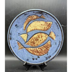 May include: A round ceramic plate with a blue background and four fish designs. The fish are tan and brown with textured scales and fins. The plate has a dark brown rim and is displayed on a black stand.