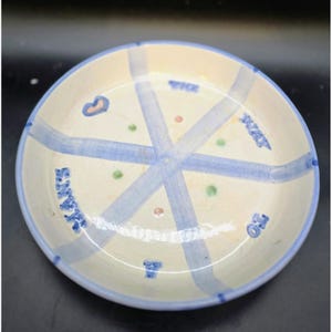 May include: A ceramic plate with a cream-colored base and a blue rim. The plate features a crisscross pattern of light blue lines, with small colorful dots and text. The word "SAY" is visible on the plate.