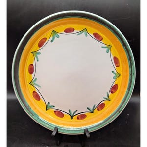 May include: A round ceramic plate with a white center, a yellow band, and a green and brown border. The yellow band is decorated with red oval shapes and green leaf-like designs. The plate is on a black stand.