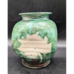 May include: A green ceramic vase with a white leaf design. The vase has a rounded body and a wide mouth.
