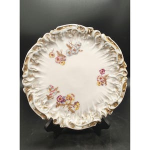 May include: A white porcelain plate with a gold rim and floral design. The plate has a scalloped edge and is decorated with pink, yellow, and blue flowers.