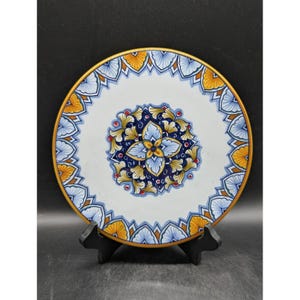 May include: A decorative ceramic plate with a white background, featuring a central floral design in blue, yellow, and red. The rim is adorned with a repeating pattern in blue and yellow, and a gold border.