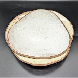 May include: Two ceramic plates with irregular edges. The top plate is a speckled off-white, and the bottom plate is a pale yellow. Both plates have a brown rim. The plates are stacked on top of each other.