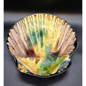 May include: A decorative, shell-shaped bowl with a glossy finish. The bowl features a vibrant color palette of yellow, green, brown, and blue. The interior has a textured, fan-like pattern, and the rim is black.