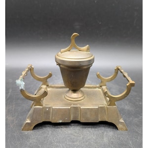 May include: Antique brass inkwell with a hinged lid and a decorative handle. The inkwell sits on a rectangular base with curved supports on each side. The brass has a warm, aged patina.