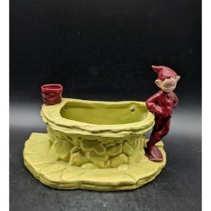 May include: A ceramic planter shaped like a stone well with a light green textured exterior and a smooth interior. A small red-clad elf figure stands on the right side of the well, leaning against the rim. A small red bucket sits on the left rim.