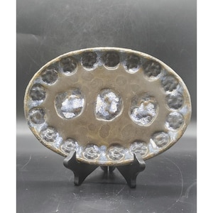May include: Oval ceramic serving dish with a dark brown and blue speckled glaze. The dish has three larger indentations in the centre and smaller circular indentations around the edge. It is displayed on a black stand.