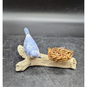 May include: A ceramic bluebird figurine perched on a faux wooden branch, with a small nest beside it. The bird has a light blue body and a darker blue tail. The nest is made of intertwined twigs. A decorative item.