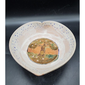 May include: A heart-shaped ceramic bowl with a white glaze and blue speckles. The bowl features a painted design of a tree with a brown trunk and green leaves, surrounded by a circle of pink flowers.