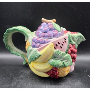 May include: A ceramic teapot with a colorful fruit design. The teapot features a lid shaped like a bunch of purple grapes, a watermelon slice, bananas, and other fruits. The handle and spout are green.