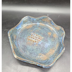 May include: A handmade ceramic dish with a wavy rim. The dish features a blend of blue and brown hues, with a textured surface. A cluster of gold-colored decorative elements is centered on the dish. This unique piece is ideal for serving or display.