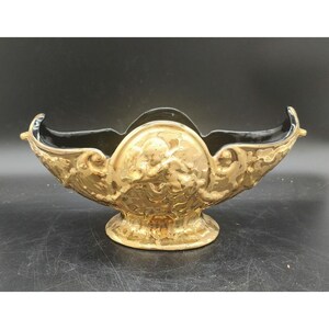 May include: A gold-colored decorative bowl with a black interior. The bowl features ornate relief detailing, including cherubic figures and scrolling patterns. The bowl has a pedestal base and curved handles on each side.