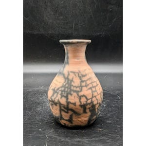 May include: A small, handmade ceramic vase with a narrow neck and a rounded body. The vase features a natural, earthy color palette with a reddish-brown base and a cracked, black pattern. The vase is likely used for decorative purposes.