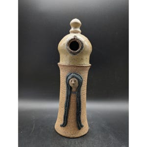 May include: A handcrafted ceramic birdhouse with a unique design. The birdhouse features a tan, cylindrical base with a sculpted face and long black hair. The top is a lighter tan color with a circular opening. The birdhouse is a decorative item.