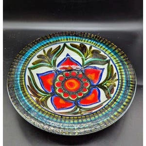 May include: A large, round ceramic bowl with a white background and a colorful floral design. The bowl has a green, blue, and brown rim.