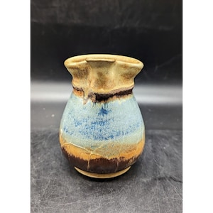 May include: A handmade ceramic pitcher with a unique, organic shape. The pitcher features a color palette of brown, tan, blue, and orange, with a dark brown band near the top. The rim has a distinctive, uneven edge.