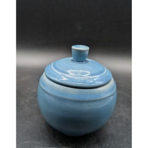 May include: A round, light blue ceramic sugar bowl with a matching lid. The bowl features a smooth, glossy finish and a small, cylindrical knob handle on top of the lid. The design is simple and spherical, with a consistent blue glaze throughout.