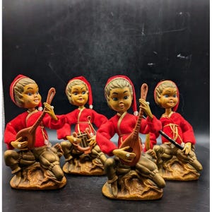 May include: Four red-clad figurines of elves playing musical instruments. The elves are sitting on wooden bases and are wearing red hats with white trim. The instruments include a lute, a lyre, a mandolin, and a flute.