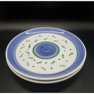 May include: Two white ceramic plates with a blue and green painted design. The plates have a blue border and a central spiral design in blue and green. Small green leaf-like shapes are scattered across the white surface.