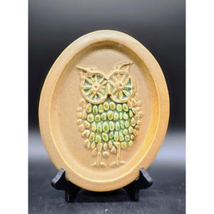 May include: Oval ceramic wall plaque with a raised owl design. The owl has green and tan details, with a textured body and large eyes. The plaque is a warm tan colour and is displayed on a black stand.