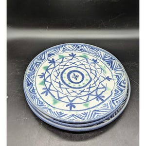 May include: A set of three ceramic plates with a blue and white geometric design. The plates feature a central circle with a starburst pattern and a border of blue and white zigzags.