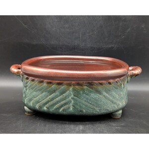 May include: A ceramic planter with a dark red rim and interior. The exterior is a teal green with a leaf pattern. The planter has two handles and three small feet. A decorative piece for plants.