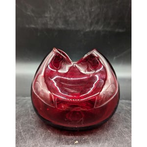 May include: A deep red, teardrop-shaped glass vase with a unique, open-mouthed design at the top. The vase has a smooth, glossy finish and sits on a dark surface. The overall shape is reminiscent of a stylized flower.