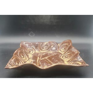 May include: A brown ceramic decorative bowl with a unique leaf shape. The bowl features a textured surface with swirling patterns and the letter "K" in the center. The edges have a wavy design, and the bowl is set against a dark background.