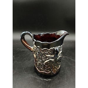 May include: A dark purple glass pitcher with a grapevine design. The pitcher has a wide mouth and a handle.