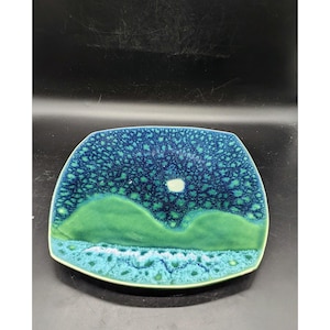 May include: A square ceramic plate with a green and blue glaze. The plate has a design of a landscape with hills and a white moon.