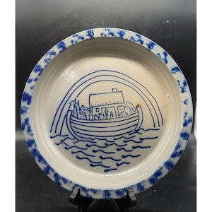 May include: A round, off-white ceramic plate with a blue rim featuring a hand-drawn illustration of Noah's Ark. The ark is depicted with animals and people under a rainbow. The plate's rim has a blue spotted pattern.