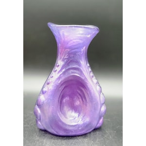 May include: A pearlescent purple vase with a flared top and a bulbous body. The vase has a textured surface with a swirling pattern and small, rounded embellishments along the sides. The color shifts in the light, creating a shimmering effect.