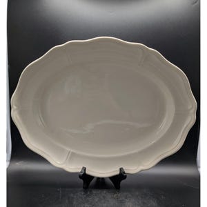 May include: An off-white, oval serving platter with a scalloped edge. The platter has a slightly raised rim and a smooth surface. The design includes subtle rectangular details along the edge. The platter is displayed on a black stand against a dark background.