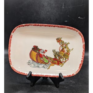 May include: Rectangular ceramic serving platter with a cream-colored surface and a red, textured border. The platter features a hand-painted illustration of Santa Claus in his sleigh, pulled by reindeer, a festive holiday theme.