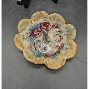 May include: A flower-shaped ceramic ring dish with a central post. The dish features a glazed surface with swirling patterns in shades of deep red, muted green, and light blue, set against a beige base. The edges are scalloped and rounded.