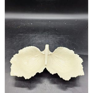 May include: A white ceramic serving dish shaped like two leaves with a handle in the center. The dish has a textured surface and is designed for serving snacks or appetizers.