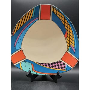 May include: A triangular decorative plate with a cream-coloured centre and a vibrant border. The border features a blue background with orange, yellow, and pink geometric patterns. The plate is displayed on a black stand.