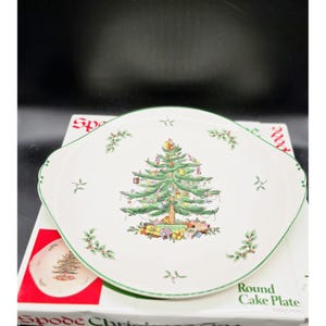 May include: White Spode Christmas Tree round cake plate with a green rim. The plate features a detailed illustration of a decorated Christmas tree with presents underneath. The plate is on top of a box with the words "Round Cake Plate".
