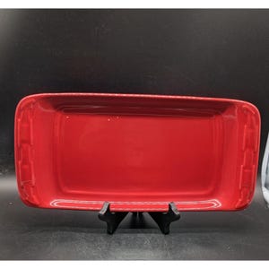 May include: A rectangular, glossy red ceramic serving tray with rounded corners and raised edges. The short ends of the tray feature a decorative, molded vertical bar pattern. The tray is supported by a black plastic display stand.