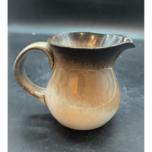 May include: A small, ceramic pitcher with a rounded body and a handle. The pitcher has a brown and tan glaze, with a darker brown rim and handle. The spout is slightly pointed. The pitcher is likely used for serving liquids.