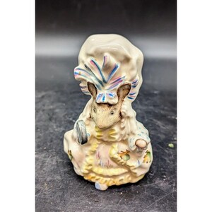 May include: A porcelain figurine of a mouse wearing a white bonnet with blue and pink trim. The mouse is holding a small, round mirror in its right paw. The figurine is decorated with floral details and has a yellow base.