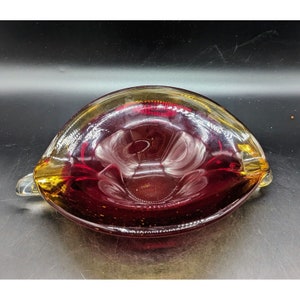 May include: A red and amber glass ashtray with a unique, wavy design. The ashtray is made of clear glass with a red interior and an amber rim.