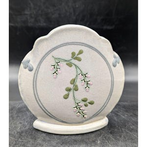 May include: A white ceramic vase with a scalloped edge and a floral design. The vase has a round base and a circular opening at the top. The floral design features pink flowers and green leaves.