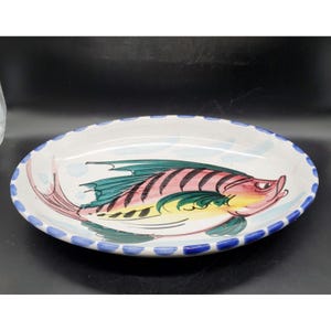 May include: A white oval ceramic serving platter with a blue and white scalloped edge. The platter features a hand-painted design of a colorful fish with red, yellow, green, and black stripes.